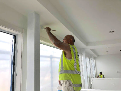 Person in a high-visibility vest painting a wall in a room with large windows.