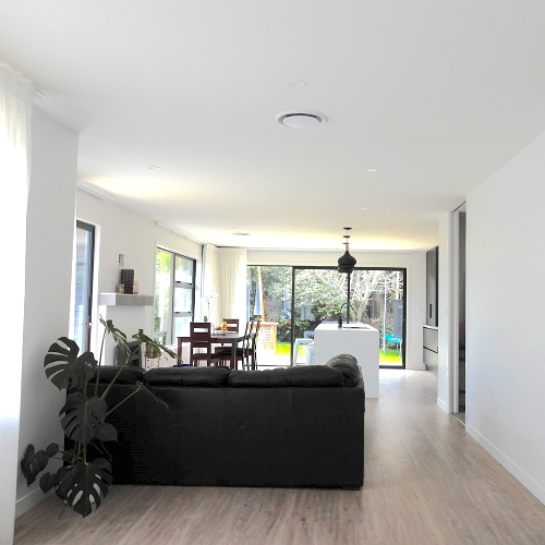 Modern living room with a black sofa, wooden floor, and large windows.